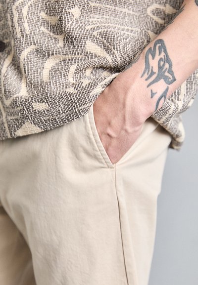 Beige-housut, joissa on sivutasku, yhdistettynä teksturoituun, abstraktikuosisesta ruskeasta ja kermanvärisestä paidasta. Lähikuva kädestä, joka lepää taskussa.