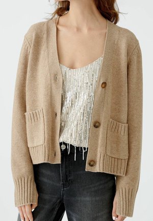 Mujer con cárdigan beige abotonado con bolsillos delanteros sobre una camiseta plateada con lentejuelas y vaqueros negros de talle alto, recortados en cuello y caderas.