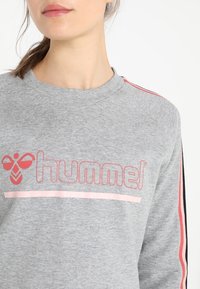 Sudadera gris con cuello redondo, que presenta un logo rosa y rayas de acento en rosa y negro a lo largo de las mangas. Textura de tejido suave.