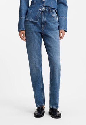 Personne portant un jean droit bleu en denim, une veste en denim assortie et des mocassins noirs, debout devant un fond blanc uni.