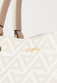 Beige mønstrets håndtaske med et geometrisk design, der har guldhardware og lysebrune håndtag. Brandlogoet vises fremtrædende.