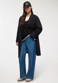 Donna che indossa un cappello nero, un lungo cappotto nero, maglie sovrapposte nere e bianche, jeans a gamba larga blu e sneakers bianche, in piedi contro uno sfondo neutro.