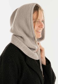 Gehaakte grijze capuchon met ribbeltextuur, gedragen over een zwarte jas. Voorzien van een trekkoord voor aanpassing en een ontspannen pasvorm rond het gezicht.