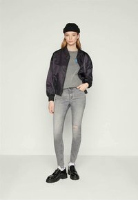 Jauna sieviete, tērpusies melnā adītā cepurē, melnā bomber jakā, pelēkā T-kreklā, saplēstās pelēkās šaurajās džinsās, baltās zeķēs un melnās masīvajās kurpēs, stāv.