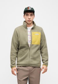 Νέος άνδρας που φοράει πράσινο φλις μπουφάν με κίτρινη τσέπη στο στήθος, πουκάμισο με σχέδια από κάτω, καφέ παντελόνι και σκούφο σκούρου πράσινου, στέκεται μπροστά σε λευκό φόντο.