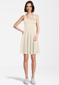 Beige mouwloze jurk met een uitlopende rok, voorzien van een Fila-logo patch op de borst. Draagt een paar grijze sneakers, van gladde stof en eenvoudig ontwerp.