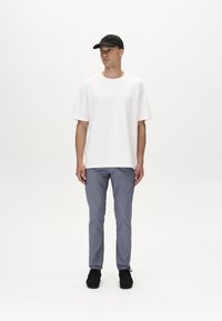 T-shirt bianca in cotone, pantaloni grigi texture, cappellino da baseball nero e scarpe nere, indossati da una figura maschile in piedi su uno sfondo bianco.