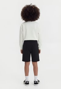 Παιδί με σγουρά μαλλιά στέκεται γυρισμένο προς τα πίσω, φορώντας λευκή μπλούζα με μακριά μανίκια, μαύρο σορτς, λευκές κάλτσες και μαύρα-άσπρα αθλητικά παπούτσια.