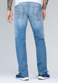 Jeans de mezclilla de color azul claro con un diseño de pierna recta, que presentan detalles de costura roja y bolsillos de parche en la parte posterior. Llevados con zapatillas.