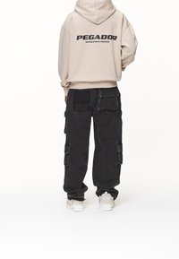 Beige luvtröja med huva och texten "PEGADOR" tryckt på ryggen, kombinerad med svarta baggy cargobyxor med flera fickor och en texturerad lapp.