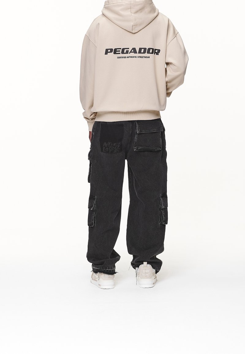 Beige luvtröja med huva och texten "PEGADOR" tryckt på ryggen, kombinerad med svarta baggy cargobyxor med flera fickor och en texturerad lapp.