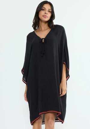 Woman wearing loose black poncho-style dress with red trim and front tie, standing against plain light background.