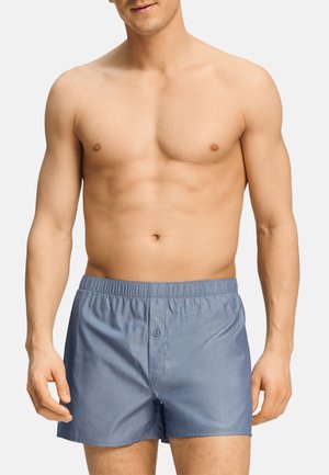 Hombre sin camiseta que lleva puestos calzoncillos tipo bóxer de algodón azul claro con cintura elástica y cierre frontal de botones sobre fondo liso.