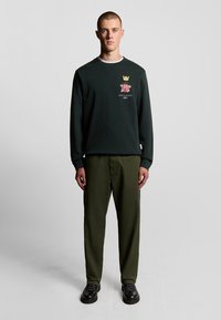 Dunkelgrüner Pullover mit einem Krone- und Rose-Logo auf der Brust, kombiniert mit olivgrünen, lässigen Hosen und schwarzen Schnürschuhen.