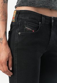 Black denim jeans featuring a slim fit, five-pocket design, and a small red and black logo label on the back pocket.