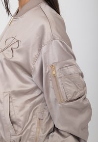 Lichtbeige satijnen bomberjack met gouden ritssluiting details en een geborduurde letter "B" op de borst, gedragen door een persoon met lang donker haar.