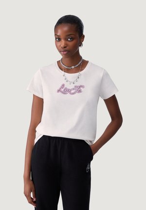 Joven mujer con cabello corto, que lleva una camiseta blanca con gráfico de Lee y pantalones negros, complementada con collares plateados en capas y pendientes de aro.