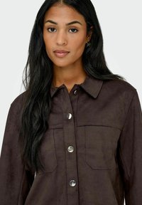 Camisa de ante marrón con cuello, cierre de botones a presión y dos bolsillos en el pecho, de textura suave.