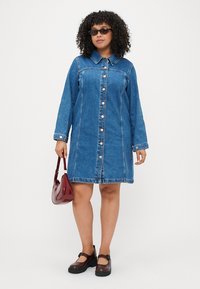 Gaiši zilā džinsa krekliņa kleita ar klasisku apkakli, priekšējiem pogām, garām piedurknēm un sānu kabatām. Pāris ar bordo rokassomu.