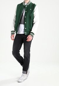 Man i grön och vit collegejacka, svarta jeans, vita högstövlar och tatueringar på händerna står framför en vit bakgrund.