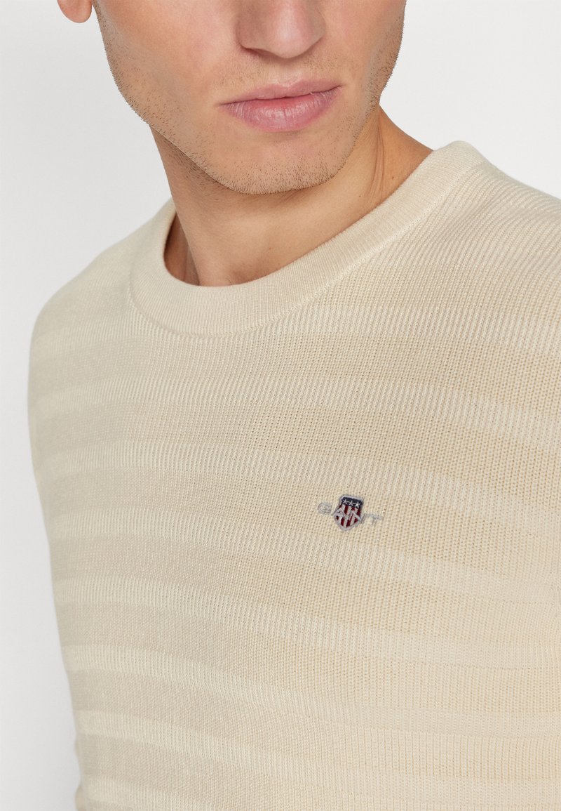Kremowy sweter z dzianiny z poziomymi paskami. Posiada okrągły dekolt i małe haftowane logo na klatce piersiowej.