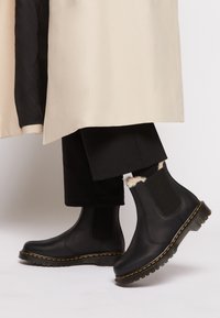 Bottes Chelsea en cuir noir avec un panneau en mesh texturé, une couture jaune le long de la semelle, et une semelle extérieure en caoutchouc épaisse. Accentuées par une fourrure en haut.