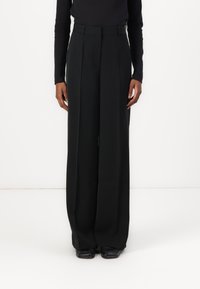 Pantalons noirs à jambe large avec une taille haute, présentant une texture lisse et des plis subtils, associés à des chaussures noires.