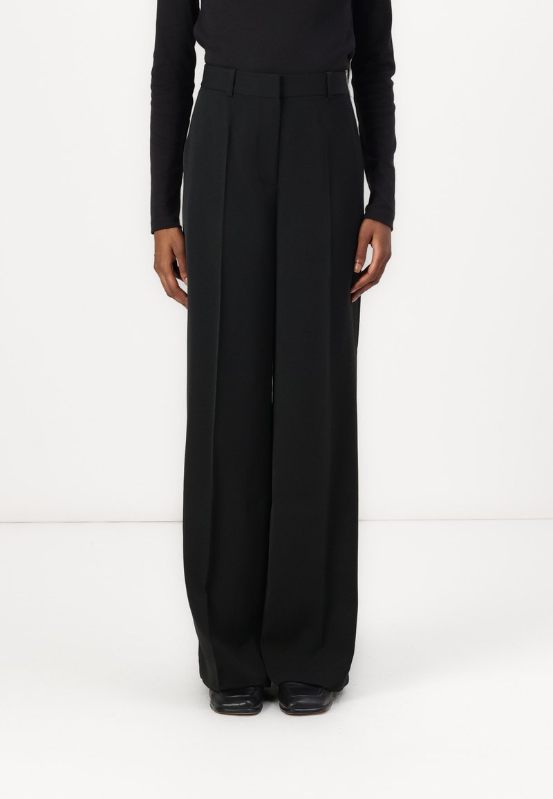 Pantalons noirs à jambe large avec une taille haute, présentant une texture lisse et des plis subtils, associés à des chaussures noires.
