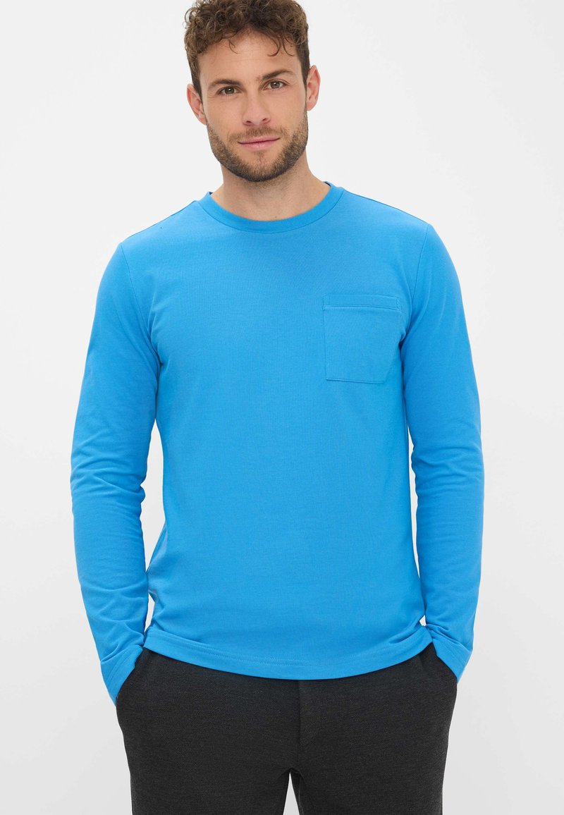 Langarm, ronde hals shirt in helderblauwe katoen met een borstzak en ribgebreide manchetten, gedragen met donkergrijze broek.