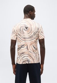 Άνδρας που φοράει π ουκάμισο με κοντά μανίκια σε μπεζ και μαύρο με μοτίβο στροβίλου και μικρό μαύρο λογότυπο στο πάνω μέρος της πλάτης, συνδυασμένο με σκούρο παντελόνι.