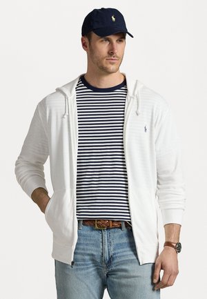 Hanorac alb cu fermoar și manșete cu nervuri, purtat peste un tricou în dungi albastre și albe, asortat cu blugi bleu deschis și o șapcă bleumarin.