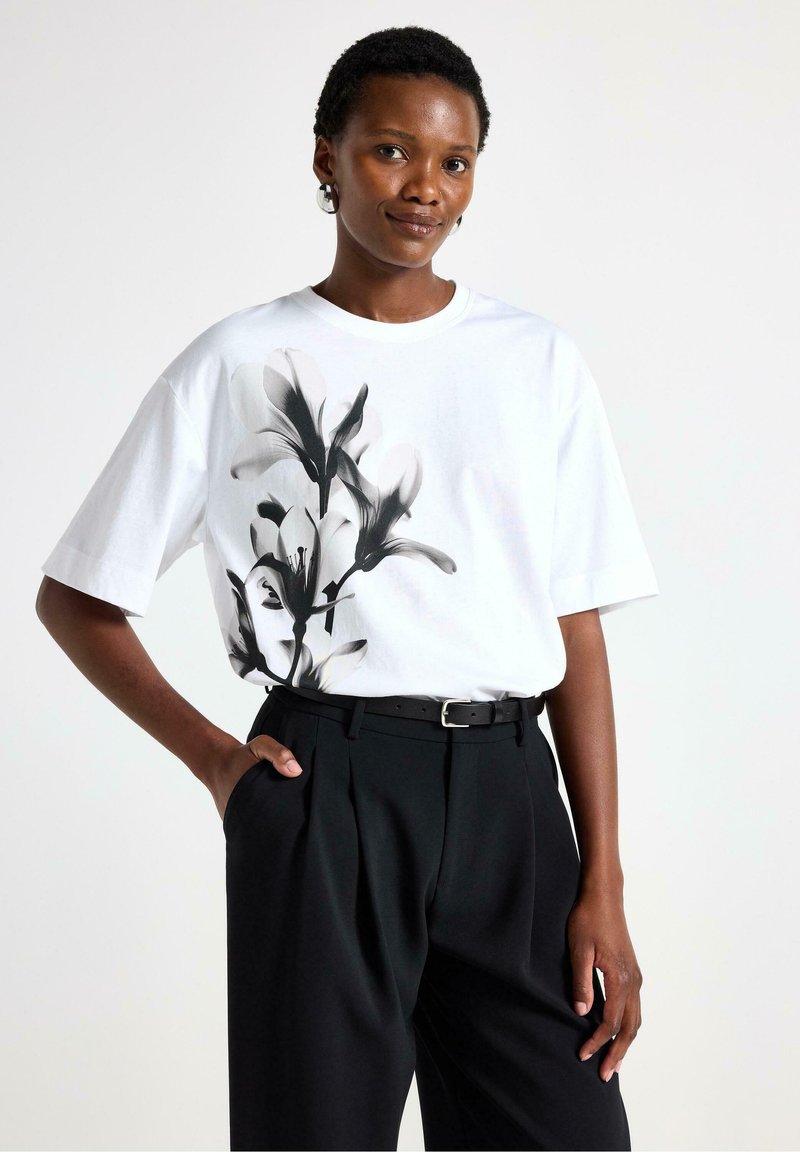 Hvid bomulds-t-shirt med sort blomsterprint på siden, kombineret med højttaljede sorte bukser og et sort bælte.