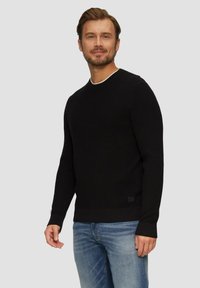 Zwarte gebreide trui met een ronde hals, een textuurstof en geribbelde boorden. G gedragen over blauwe denim jeans. Minimale merkaccent bij de zoom.