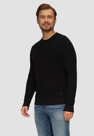 Czarny sweter dzianinowy z okrągłym dekoltem, teksturowana tkanina i prążkowane mankiety. Nosi się go na niebieskich dżinsach. Minimalistyczny akcent z logo na dole.