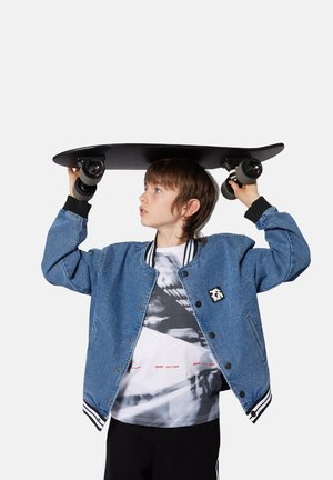 Denim bomberjack met zwarte geribde boorden, een wit en zwart graphic T-shirt, en een zwart skateboard boven het hoofd gehouden, wat een gladde textuur toont.