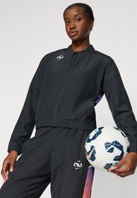 Μαύρο αθλητικό τζάκετ με φερμουάρ, ελαστικές μανσέτες και ανοιχτό ποδόγυρο, με λογότυπο στο στήθος και πολύχρωμες εναλλαγές στην σχεδίαση των μανικιών. Κρατάει μια μπάλα ποδοσφαίρου.