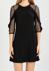 Svart klänning med trekvarts genomskinliga prickiga ärmar, med rynkad halsringning och slätt tyg. Rakt snitt, knälång design.