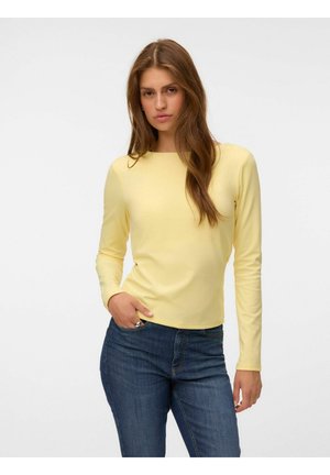 Femme aux longs cheveux bruns portant une chemise jaune clair à manches longues et un jean bleu, une main dans la poche, sur un fond uni.