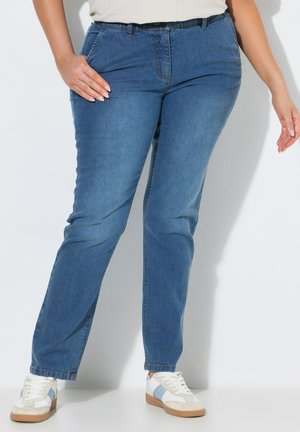 Person trägt blaue Jeans und weiße Sneaker mit hellblauen Streifen, steht mit einer Hand in der Tasche und der anderen seitlich am Körper.