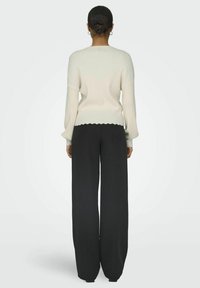 Maglione a coste crema con maniche lunghe e orlo a scallop, abbinato a pantaloni a gamba larga neri, visto da dietro.