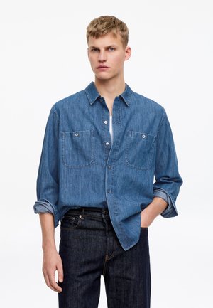 Young man with short blond hair wearing a blue denim button-up shirt over a white top and dark denim jeans, standing against white background.