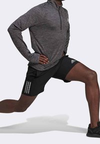 Haut de sport gris à manches longues avec un col, assorti à un short noir avec trois bandes verticales. Le mannequin montre une position de fente.