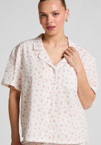 Vrouw in een wit kortmouw overhemd met een klein roze bloemenpatroon, die de kraag met haar rechterhand vasthoudt.