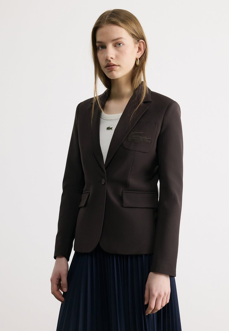 Jeune femme aux longs cheveux portant un blazer noir sur mesure, un haut blanc avec un petit logo de crocodile, des boucles d'oreilles dorées en forme de créole et une jupe plissée foncée.