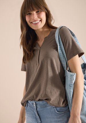 Femme souriante aux cheveux bruns portant un haut marron à manches courtes, un jean bleu rayé, et portant un sac fourre-tout en denim bleu clair sur l'épaule.