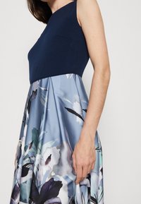 Blaues Blumenkleid mit einem taillierten marineblauen Oberteil und einem ausgestellten Rock, der große Aquarellblumen in Blau-, Grün- und Schwarztönen zeigt.