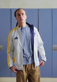 Chaqueta atlética blanca y azul marino con rayas amarillas, llevada sobre una camiseta de rayas gris. Pantalones cargo color arena. Fondo de gimnasio con taquillas visibles.