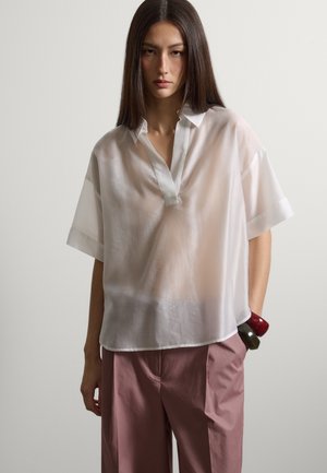 Frau mit glattem dunklem Haar trägt eine durchscheinende weiße Bluse mit kurzen Ärmeln, mauvefarbene weit geschnittene Hosen und klobige rote und grüne Armreifen am Handgelenk.