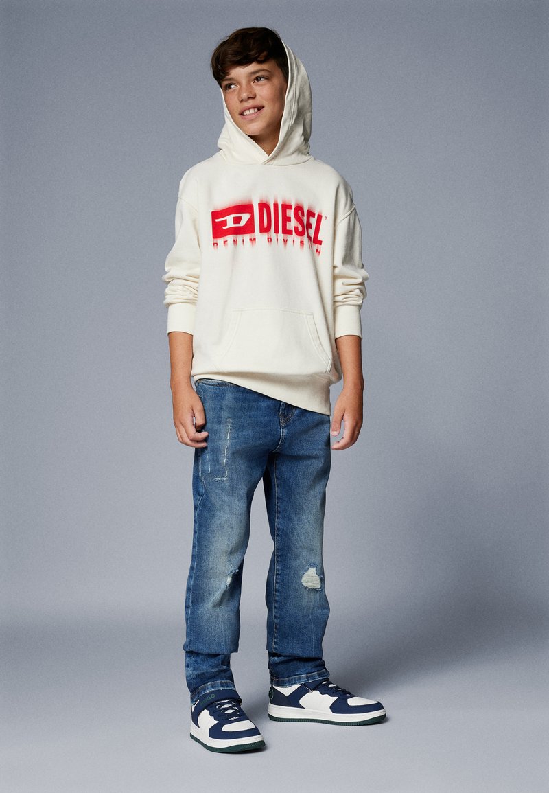 Felpa bianca con cappuccio e logo rosso "DIESEL", abbinata a jeans blu strappati e sneakers blu e bianche. Outfit semplice e casual.