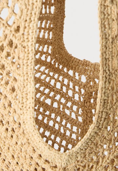 Gros plan d’un sac textile tissé beige avec un motif à grille ouverte et un détail de cadre rectangulaire arrondi.